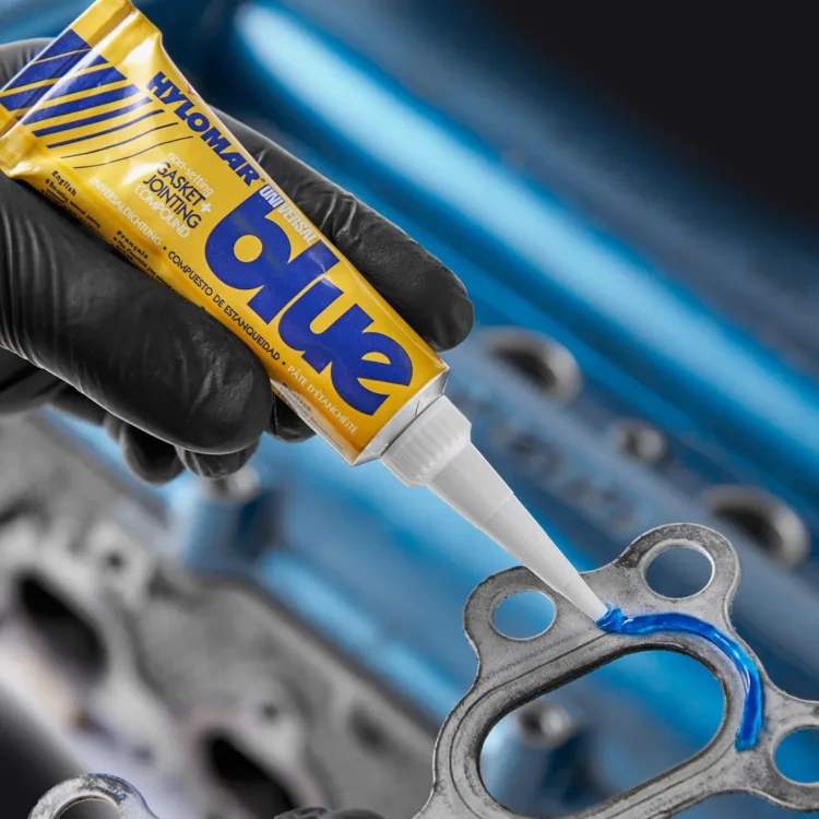 Mao com luva preta aplicando o composto de vedacao Hylomar Blue sobre uma junta metalica de motor.
