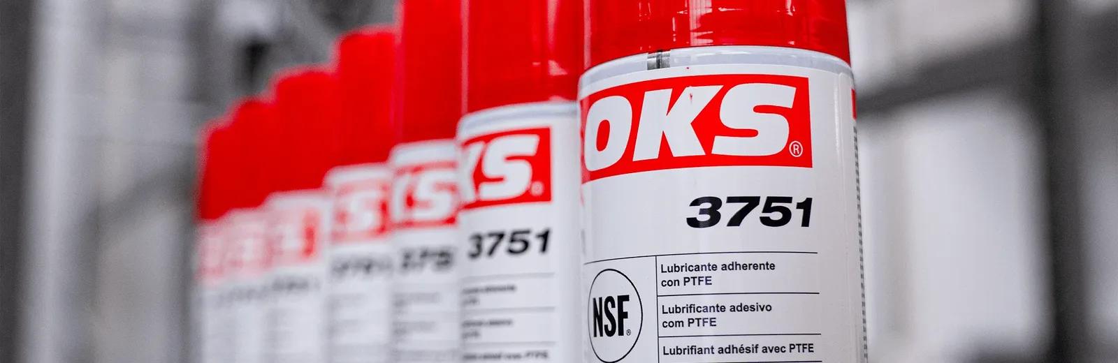 Fileira de latas spray OKS 3751, lubrificante adesivo com PTFE, em destaque sobre fundo industrial.