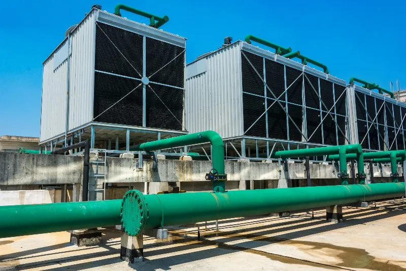 Conjunto de torres de resfriamento industriais com tubulações verdes sob céu azul em planta de refrigeração.